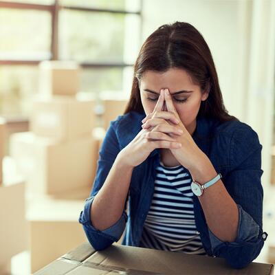 A stressed woman clasps her hands over her face while moving boxes loom in the background