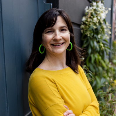 Susannah Fisher headshot. White woman with medium length dark brown hair, standing outside. She is wearing a yellow sweater and bright green hoop earrings.