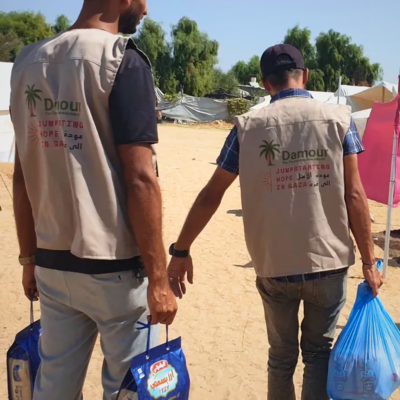 two men walking away from the camera, carrying blue bags, wearing vests that say "Jumpstarting Hope in Gaza"